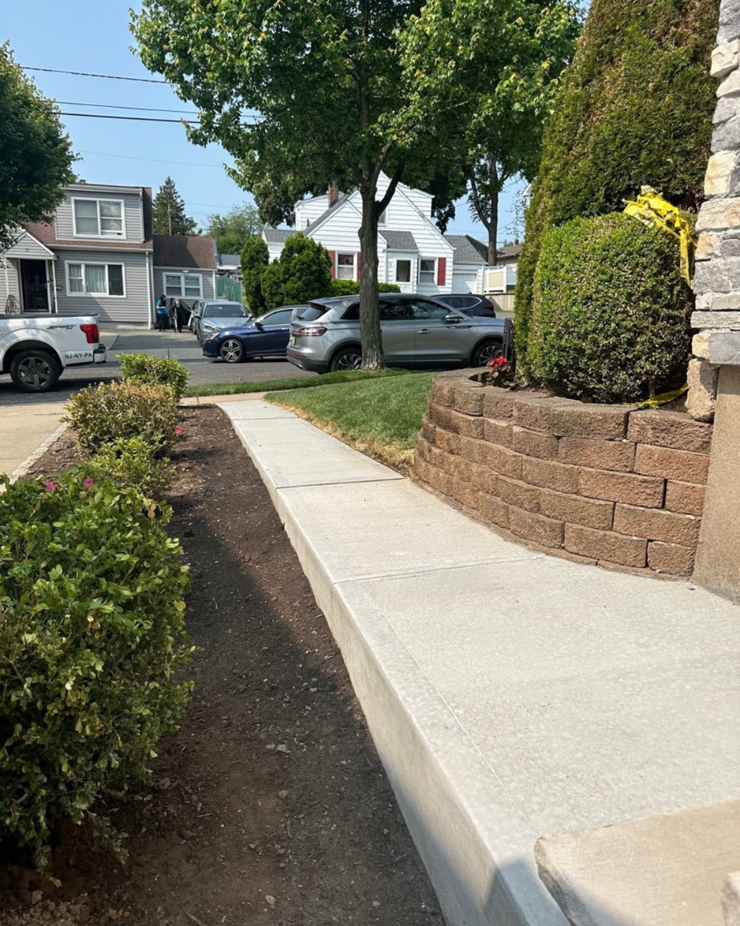 A concrete walkway bordered by plants and a retaining wall, leading to a home; street with parked cars visible.