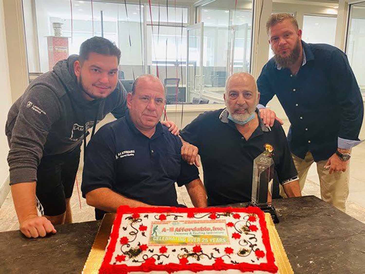 Four men around a cake, celebrating. One seated in blue, others in gray, black, and blue. Red-decorated cake.