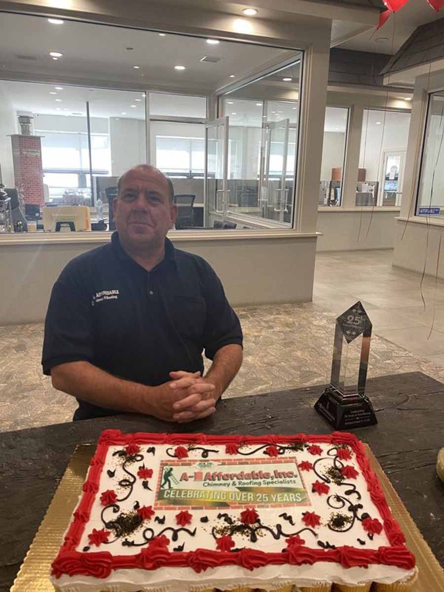 Man in black shirt sits near cake and trophy in an office setting.