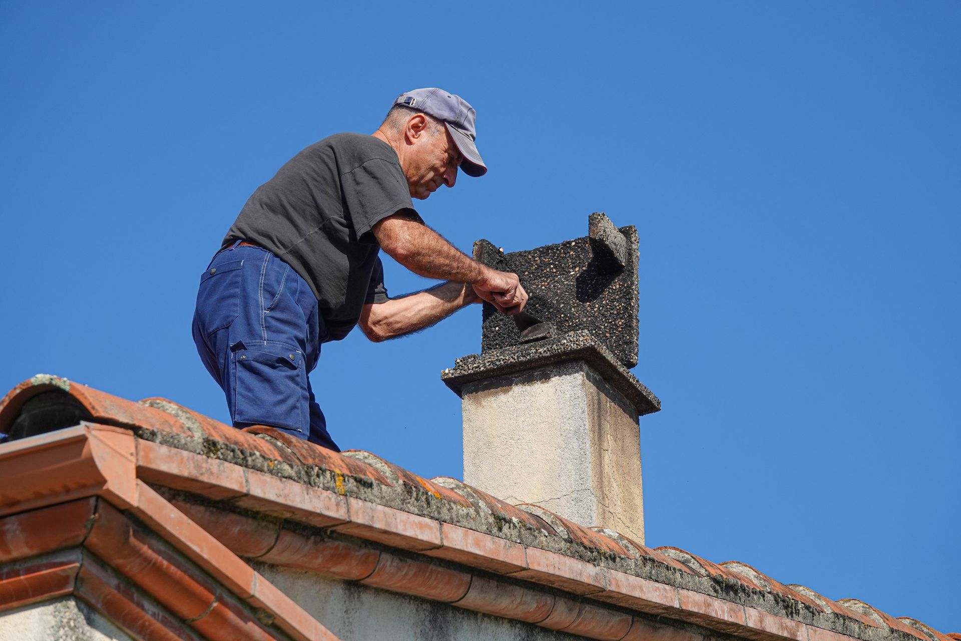 A man repairs a chimney wile standing on a house’s roof under the sun. A man repairs a chimney wile standing on a house’s roof under the sun.