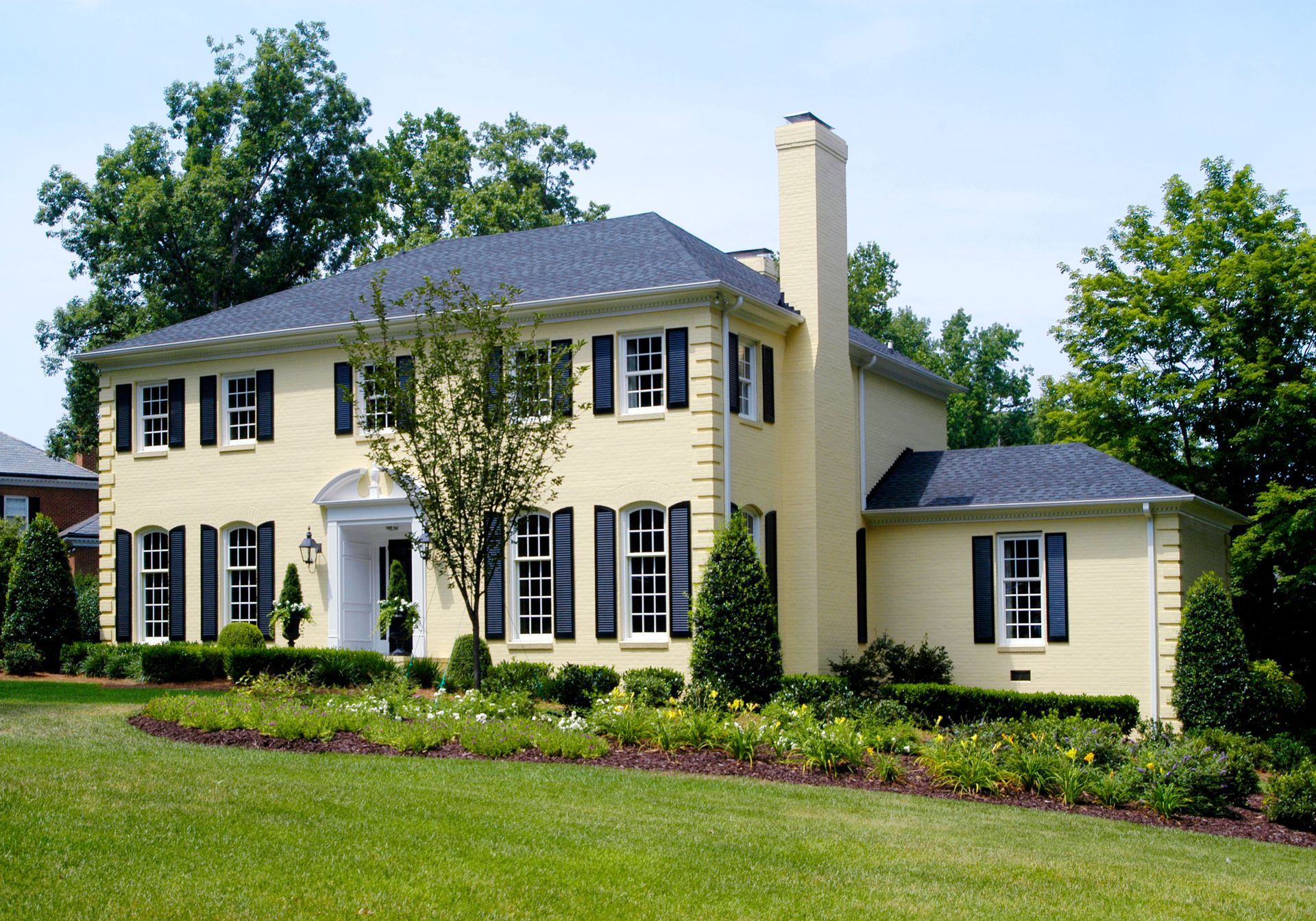Yellow two-story house with black shutters, green lawn, and landscaping under a blue sky.
