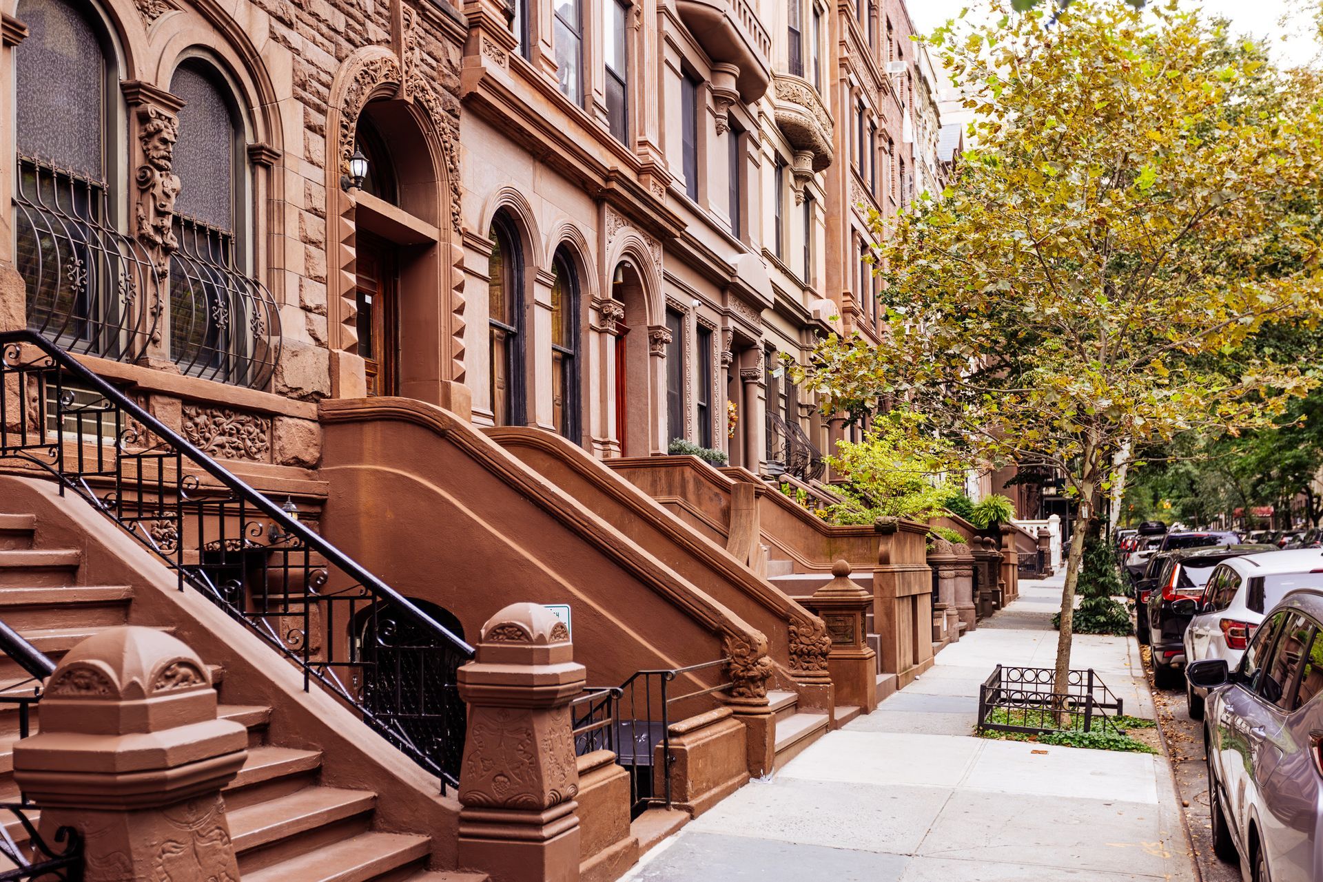 A row of brown brick buildings with stairs and cars parked on the sidewalk.