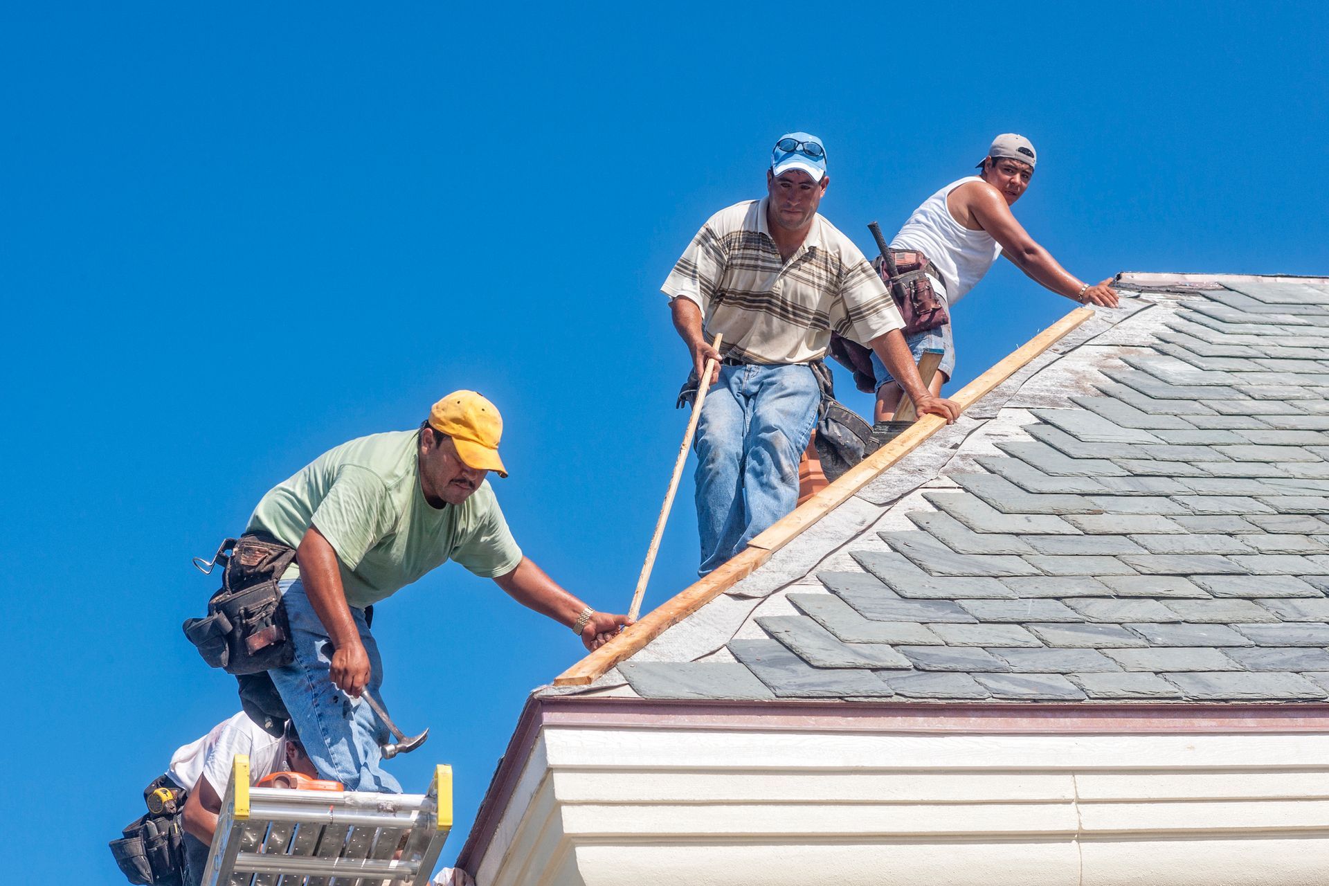 Three workers installing shingles on a house roof under clear blue sky.