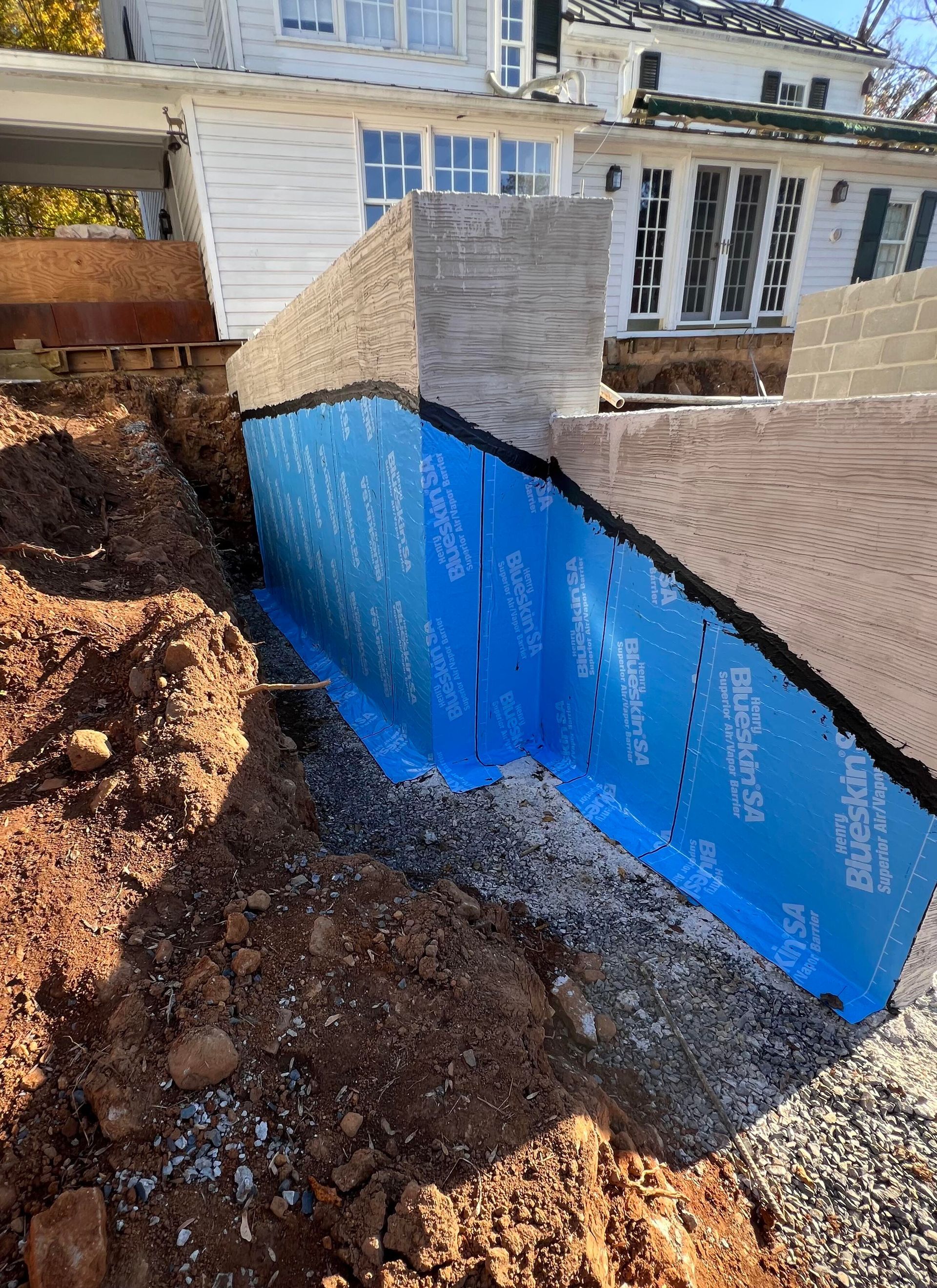 Foundation wall repair with blue waterproofing membrane, dirt and gravel in a residential setting.