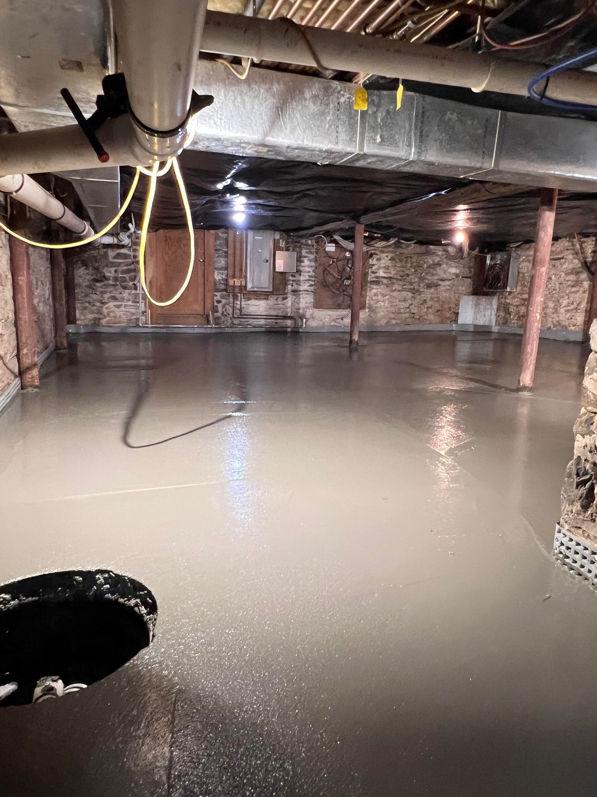 Newly poured concrete basement floor. Gray, reflective surface with pipes and brick walls overhead.