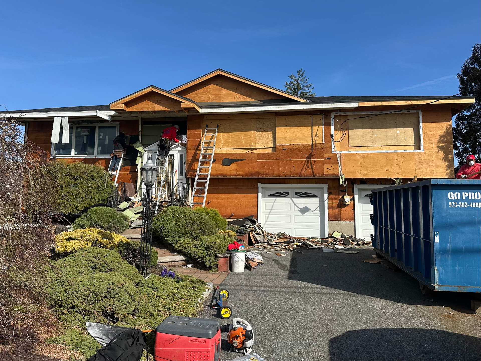 House undergoing renovation; plywood covers exposed walls. Blue dumpster, ladder, tools, and debris visible.