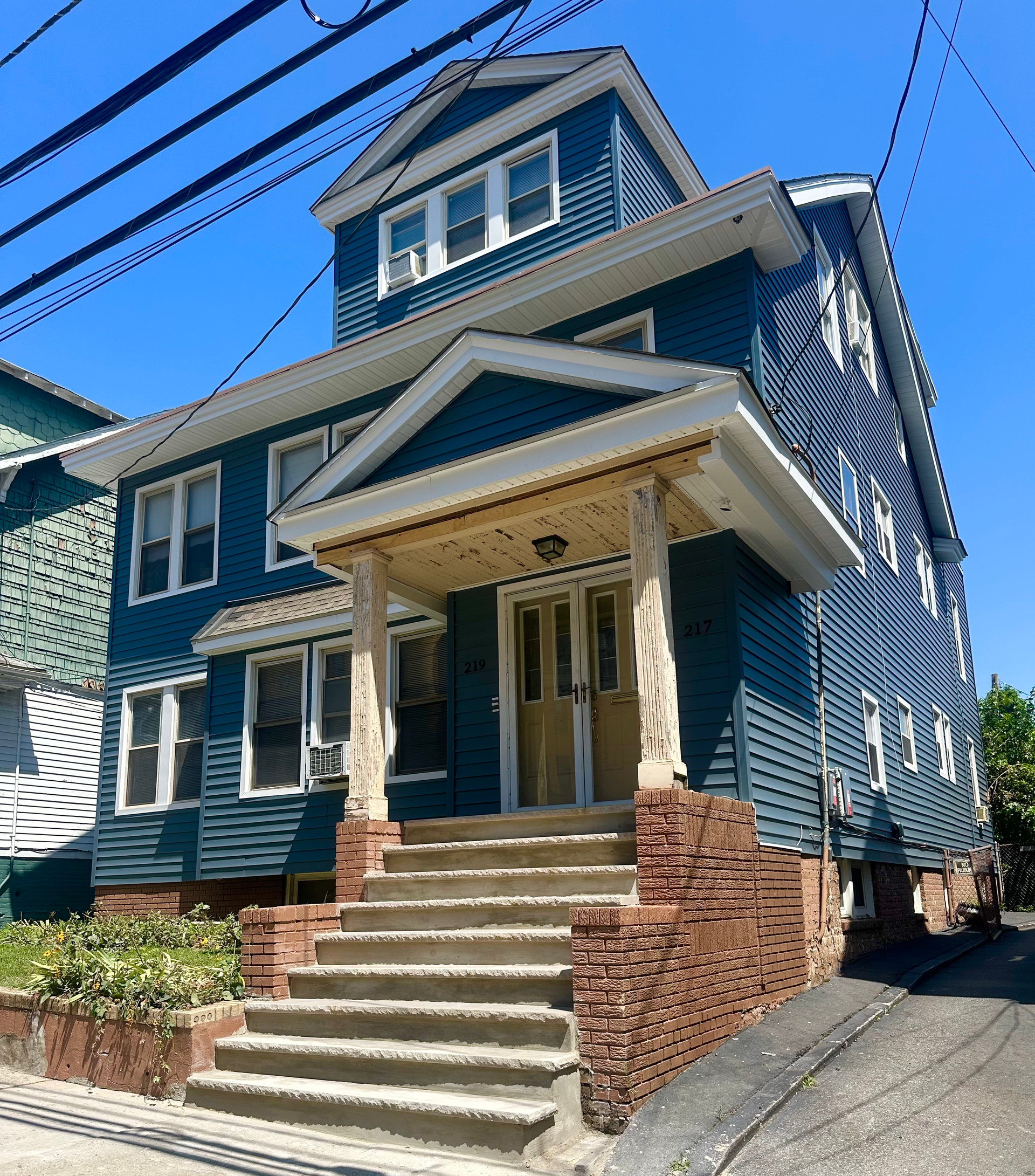 Blue multi-story house with a porch and steps, brick foundation, and clear blue sky.