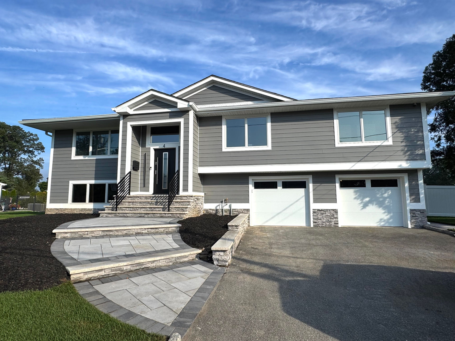 Gray two-story house with stone accents, white garage doors, and landscaped walkway on a sunny day.