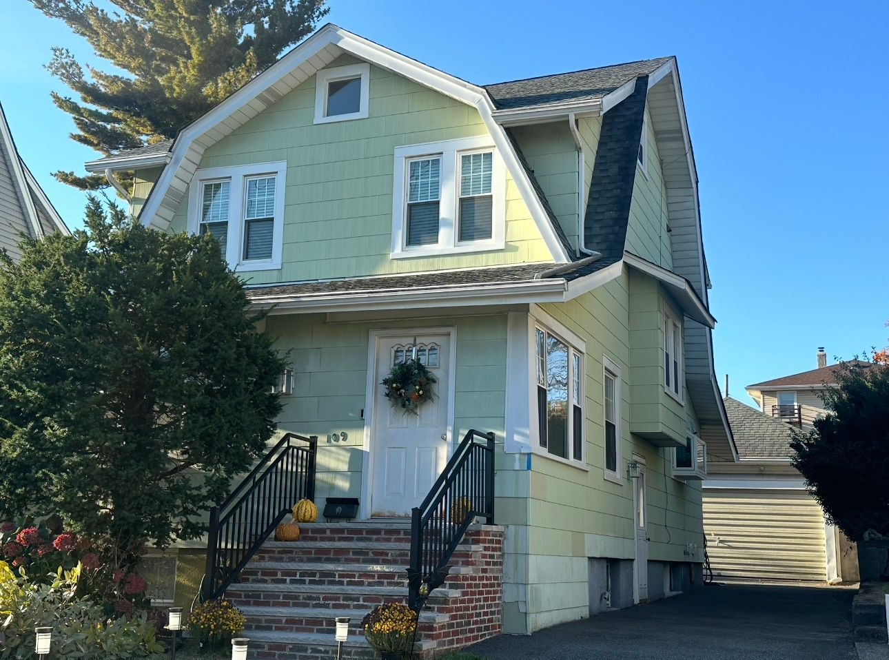 Two-story light green house with white trim, black railing, and a garage on a sunny day.