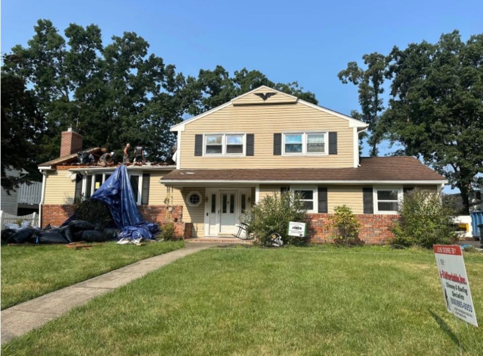 House under renovation; roofers on roof, blue tarp covers debris, yard with sign.