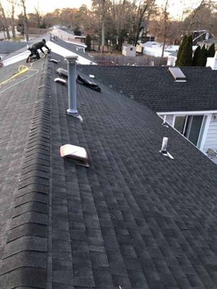 Person on a dark shingle roof with vents and a skylight; suburban setting.
