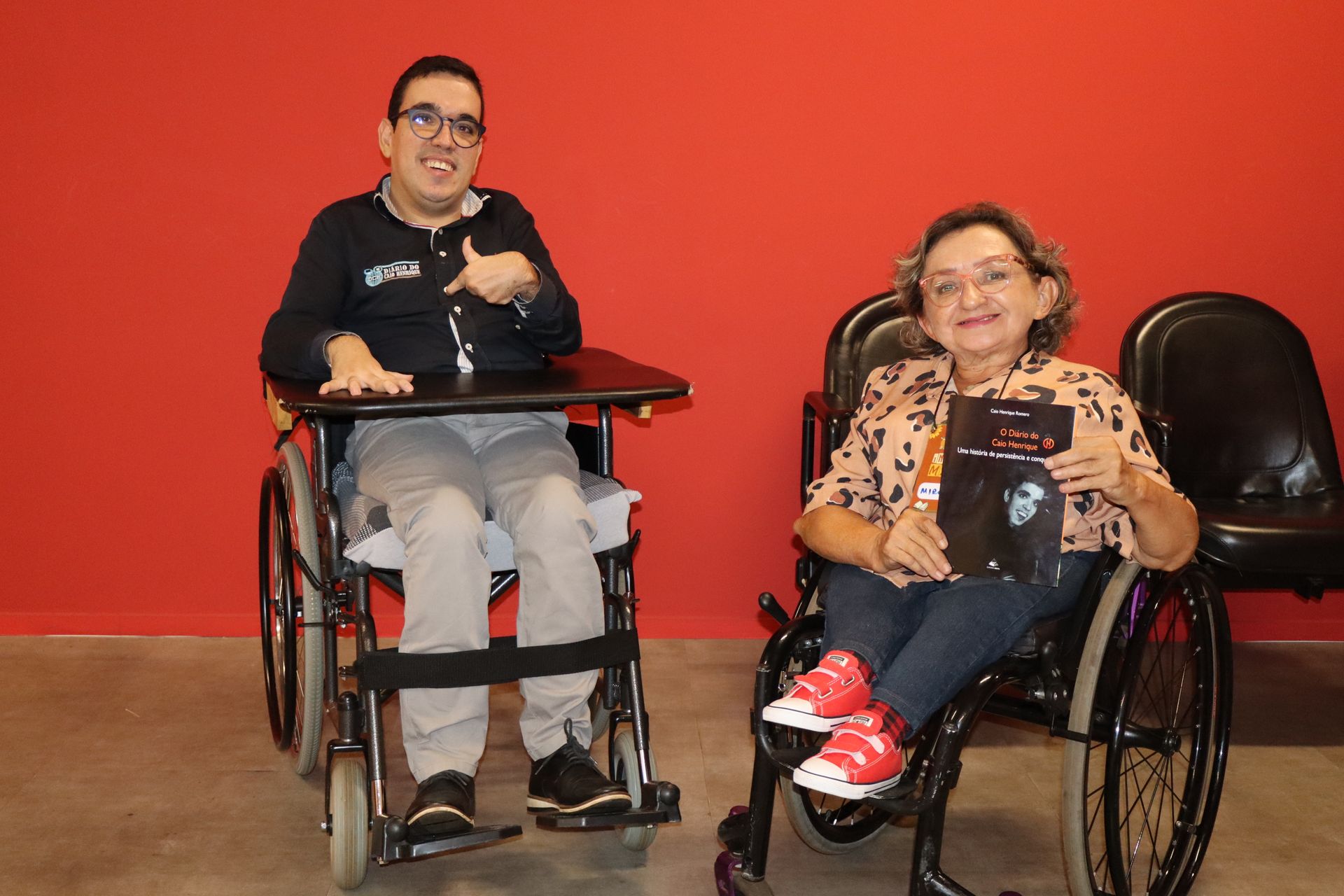 Duas pessoas em cadeiras de rodas sorrindo, uma apontando, contra uma parede vermelha. Uma delas segura um livro.