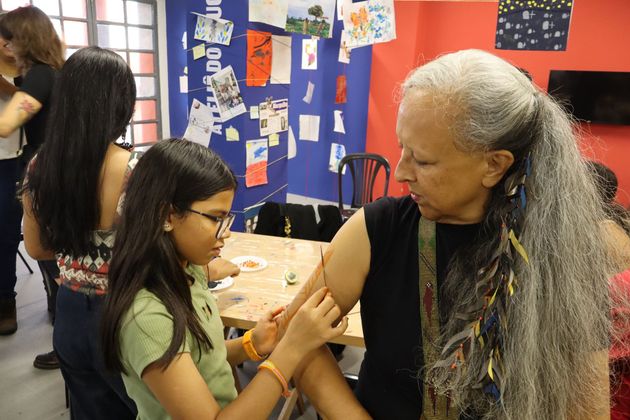 Uma criança faz pintura em uma pessoa idosa de longos cabelos grisalhos em uma sala com obras de arte na parede.