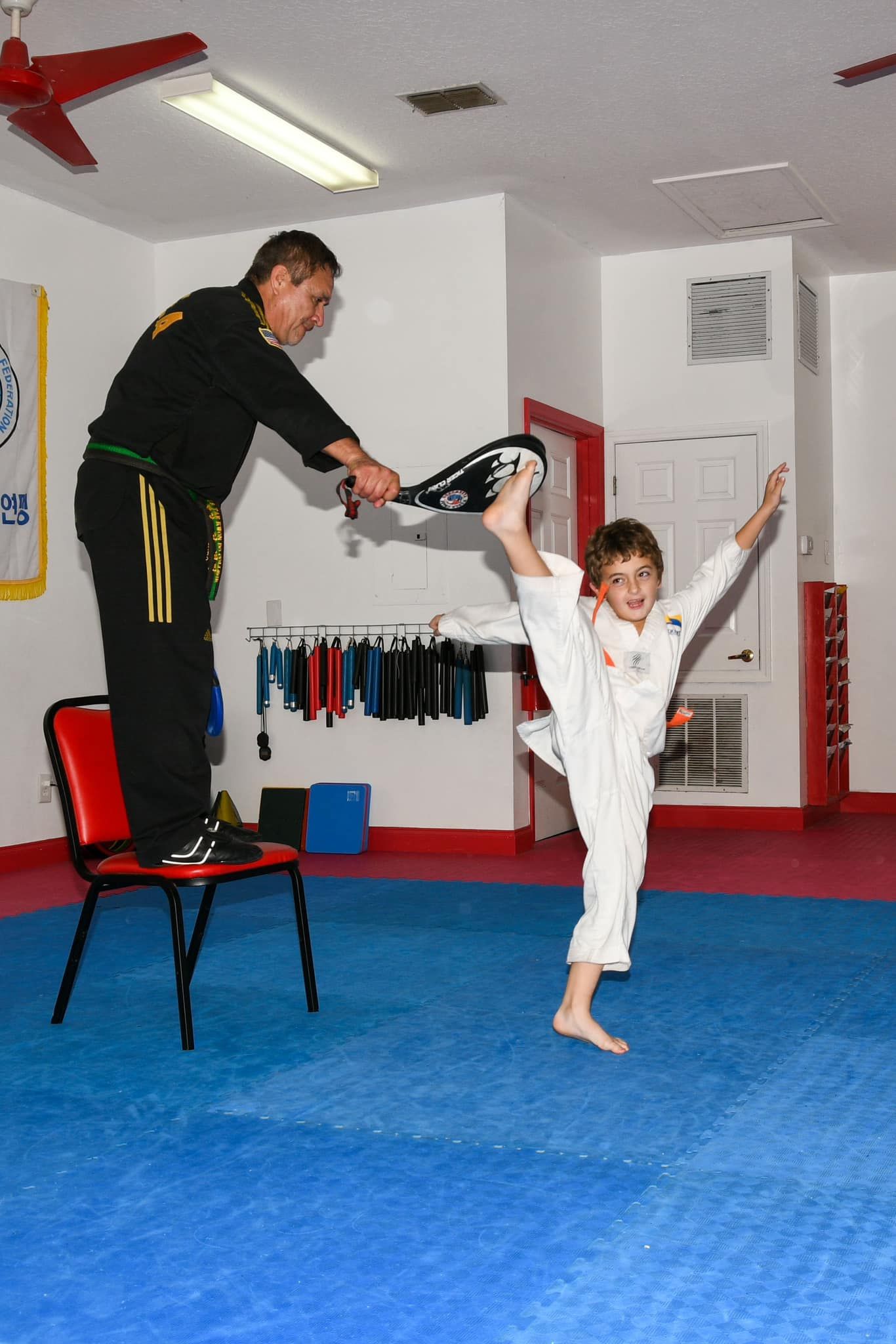 A martial arts instructor stands on a red chair holding a black pad for a student performing a high vertical kick.