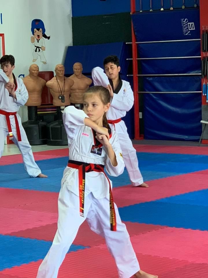 Three students in white taekwondo uniforms and red belts practice poses on a colorful mat in a gym.
