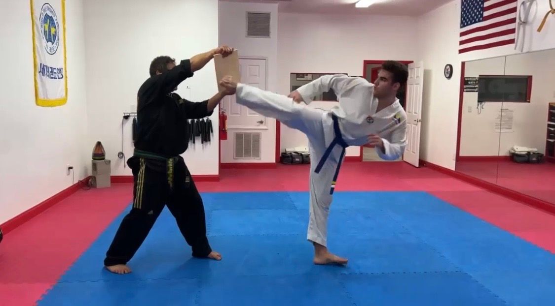 Students in white karate uniforms practice high kicks on a blue and red mat in a martial arts studio.