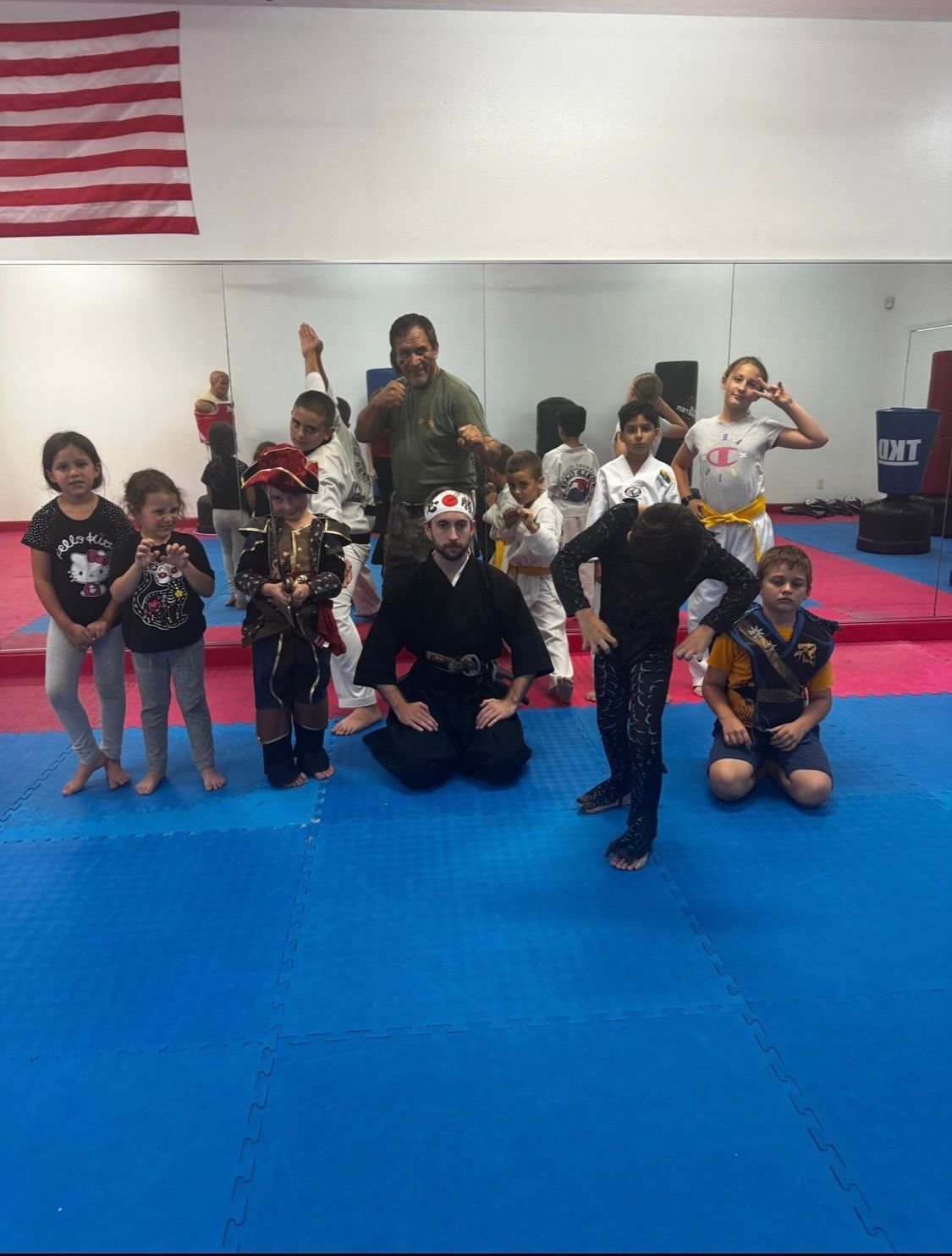 Martial arts students in white uniforms practicing stretching and warm-ups on a blue and pink mat-covered training floor.