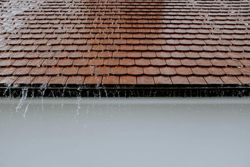 Rain cascading off a brown tiled roof into a white gutter on a building.