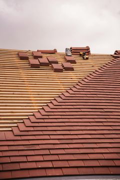 Roof construction: Half-finished roof with red tiles laid, exposing wooden beams, under a cloudy sky.