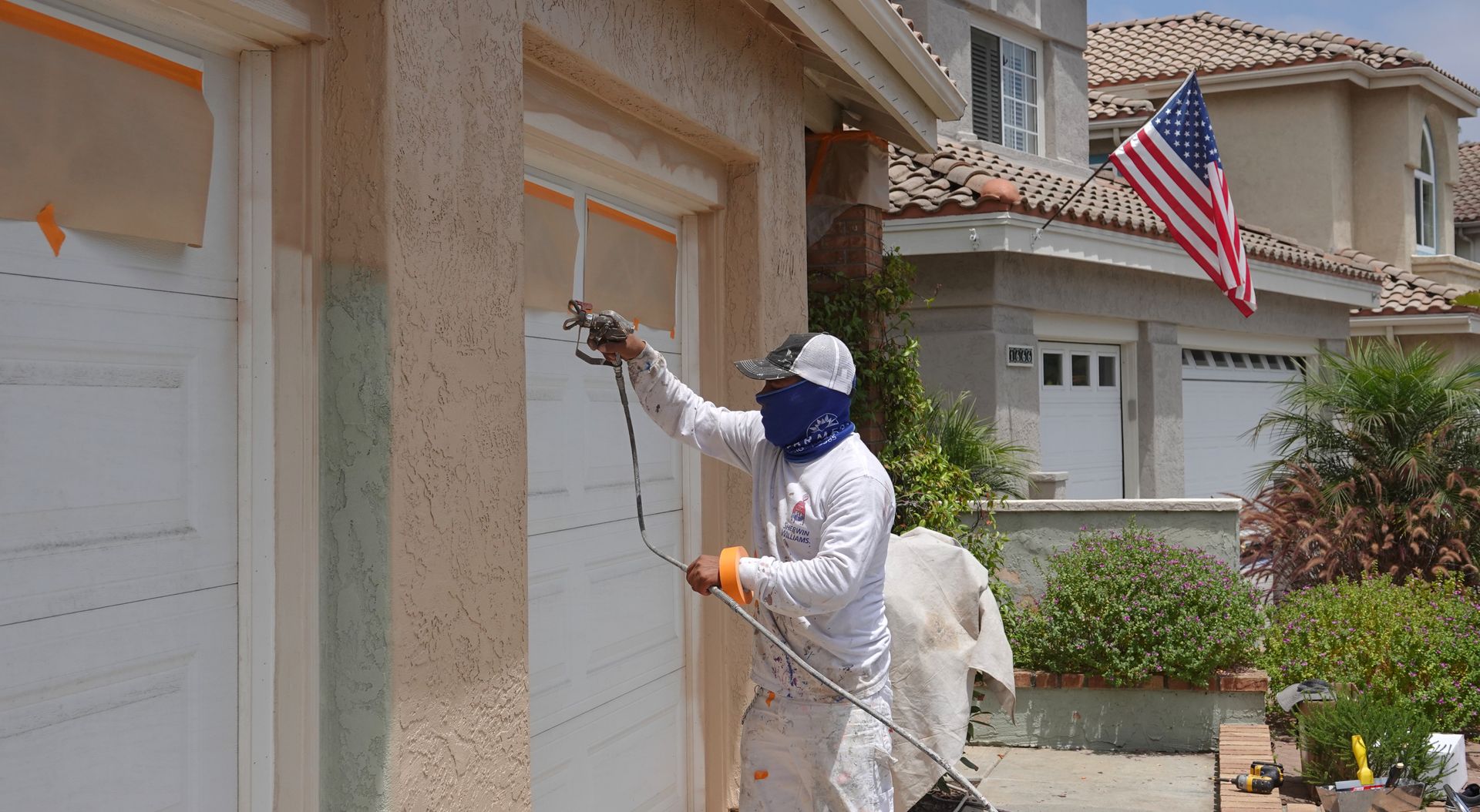 Person spray-painting a white garage door, wearing protective gear, with a house and American flag in the background.