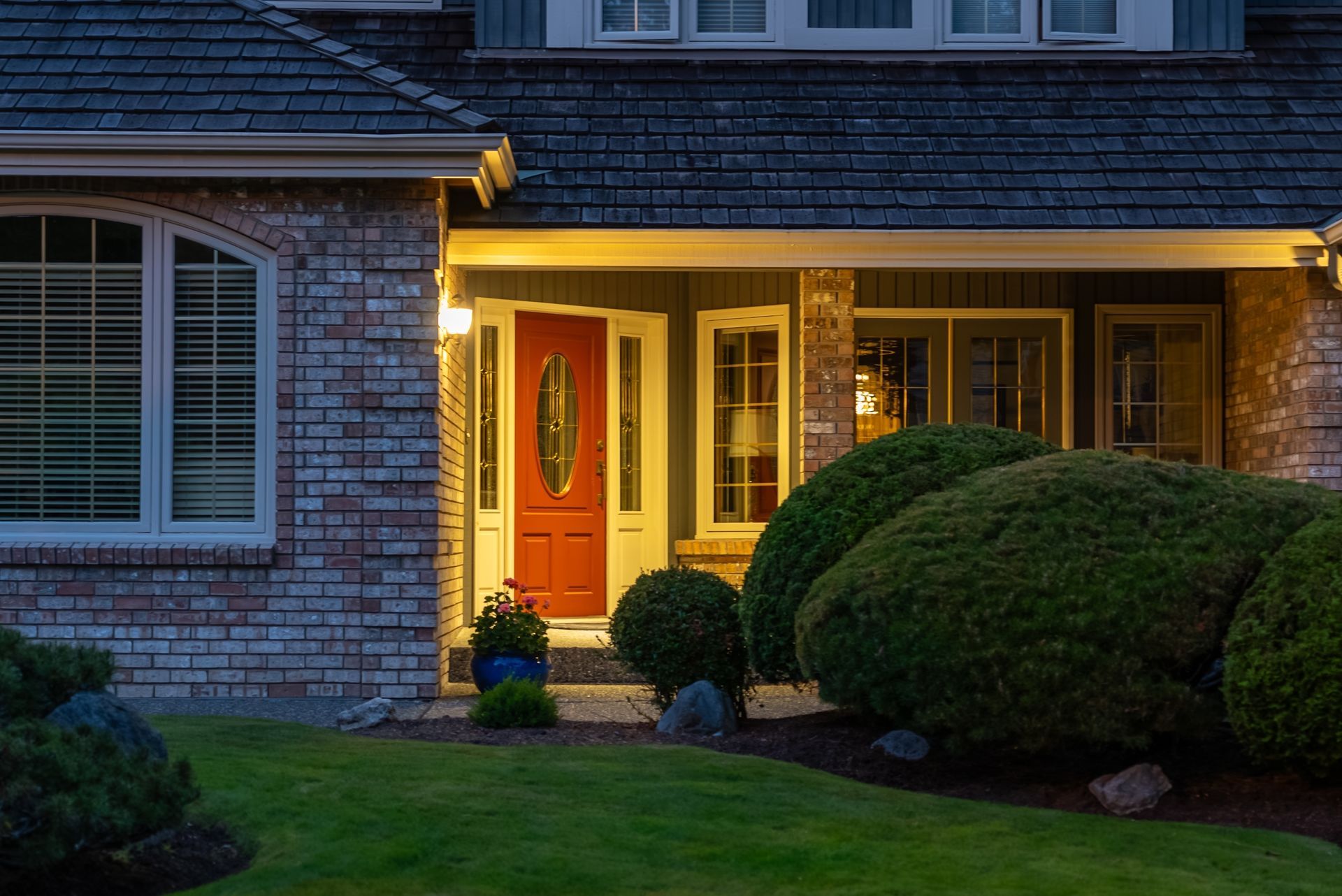 Red front door of brick house at dusk, lit by warm light. Lush green bushes and grass in foreground.
