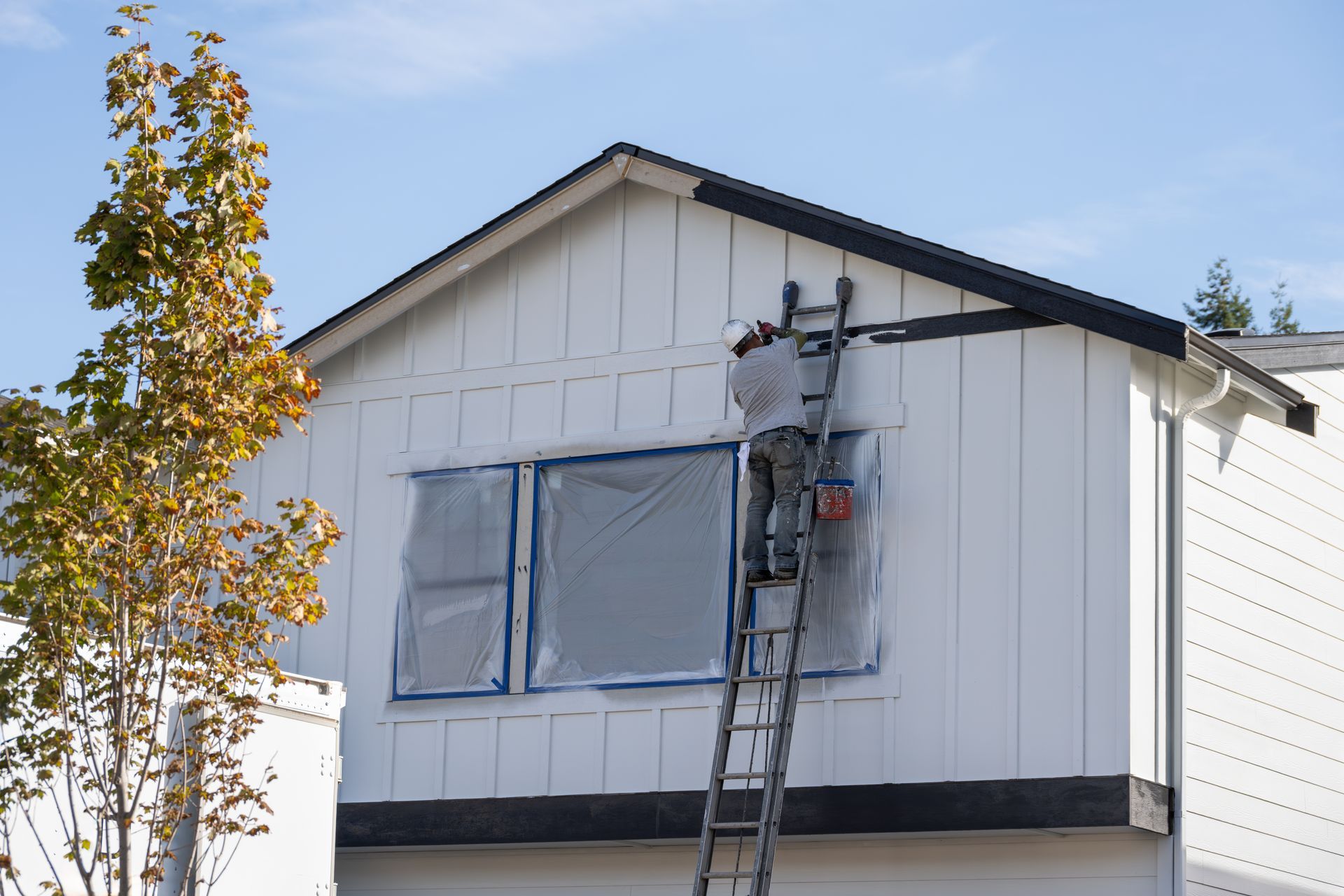 Person painting exterior of white house from ladder; windows taped off.