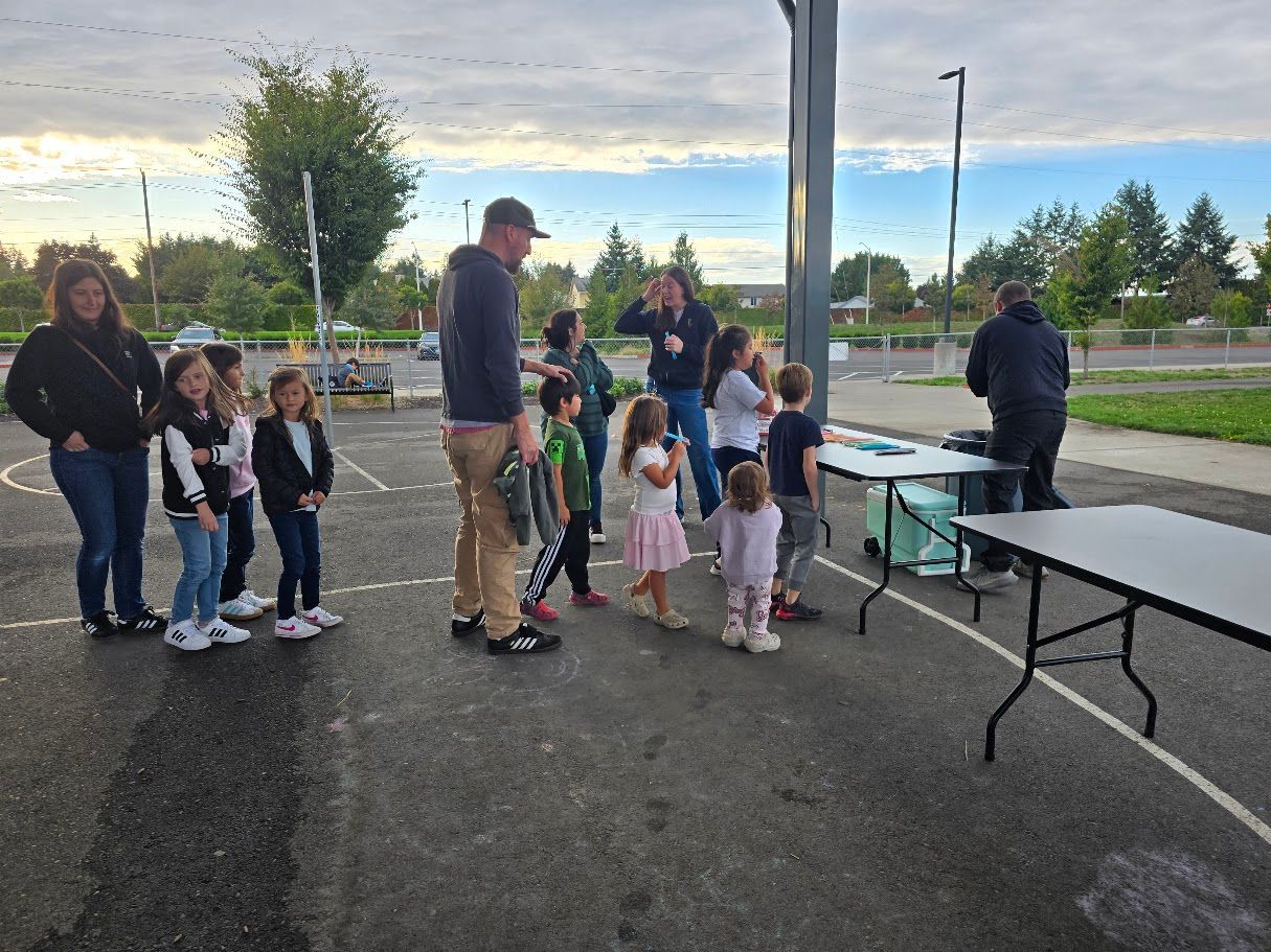 Children and adults at outdoor event; group of kids near tables, others watching. Cloudy sky.