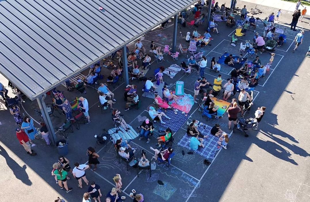 Large crowd gathered under a pavilion, sitting on chairs and blankets. Sunny day, shadows visible.