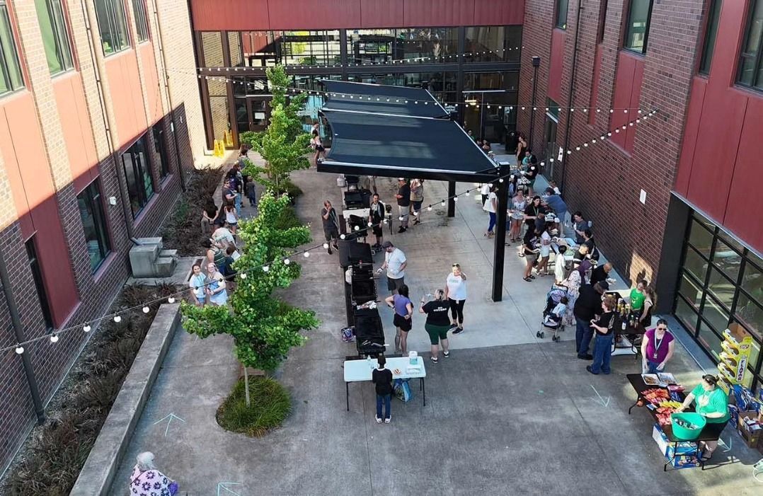 People gather in a courtyard with tables and a canopy, surrounded by red brick buildings.