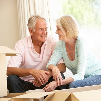Senior couple smiling, sitting together, surrounded by moving boxes, in a brightly lit room.