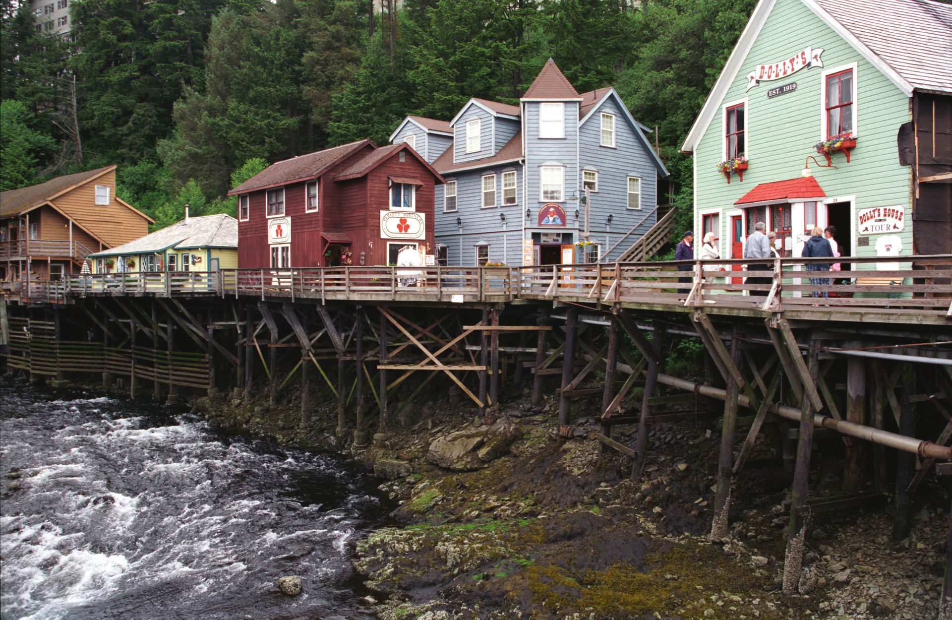Colorful buildings on a wooden pier over a river. Tourists stand outside a shop with red awning.