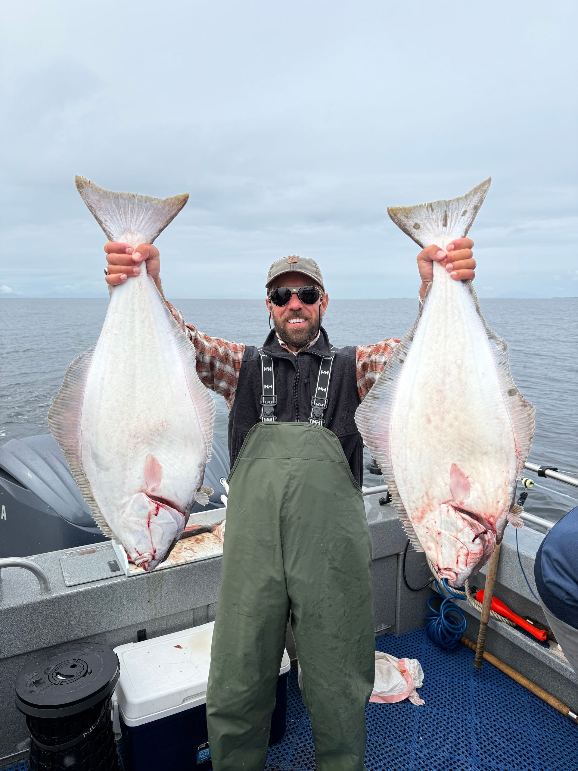 Man on boat holds up two large, flat fish (halibut) he caught in overcast water.