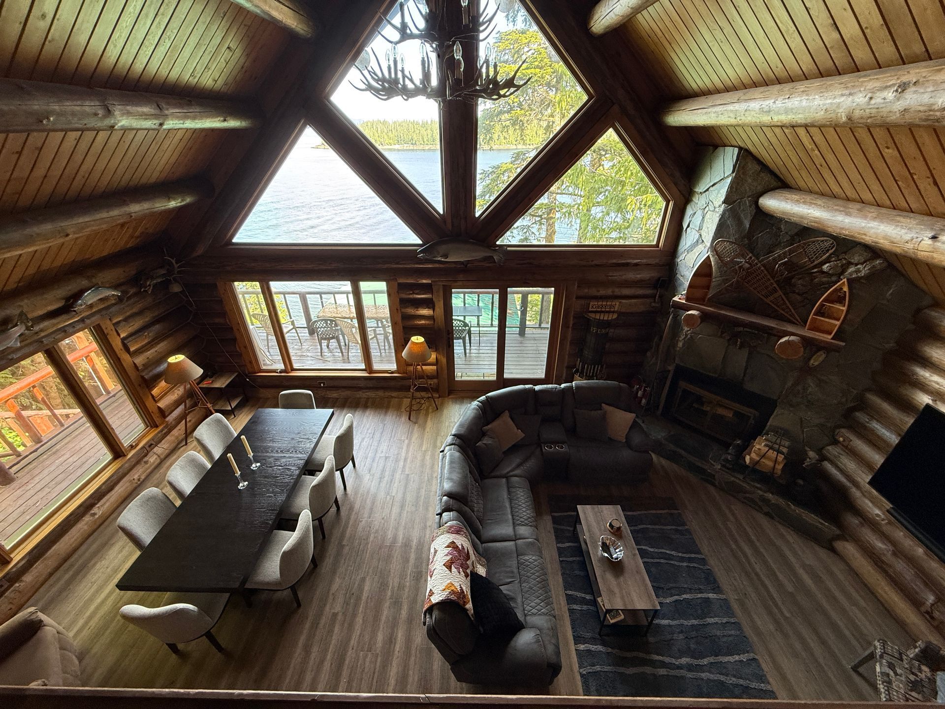 High-angle interior shot of log cabin living space with large windows overlooking water and a dining area.