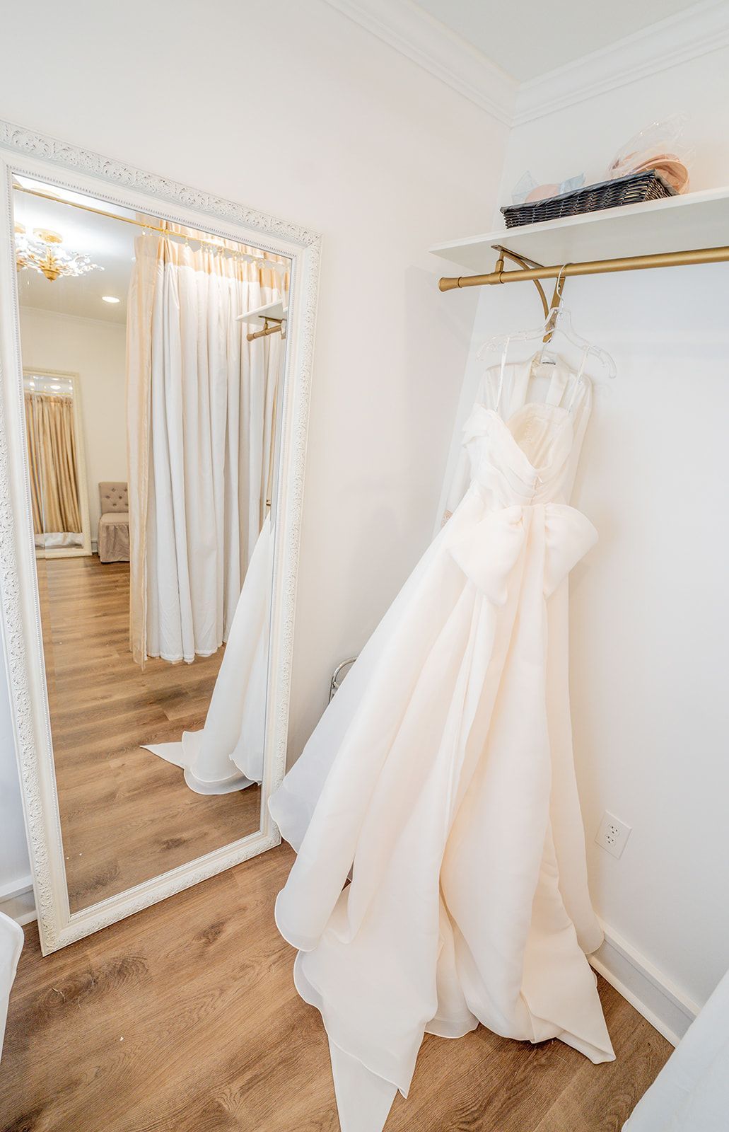 A wedding dress is hanging in a dressing room in front of a mirror.