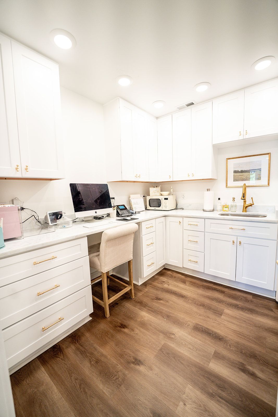 A kitchen with white cabinets and hardwood floors and a desk with a computer on it.