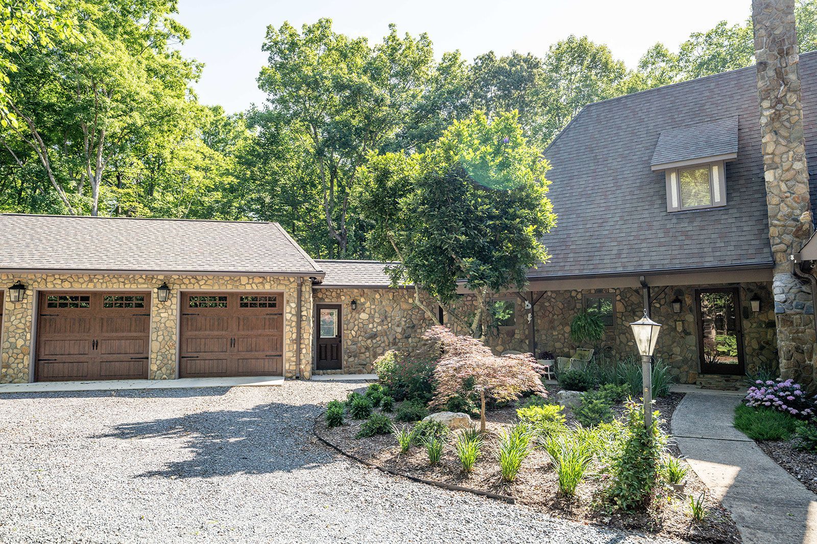 A large stone house with two garages and a driveway surrounded by trees.