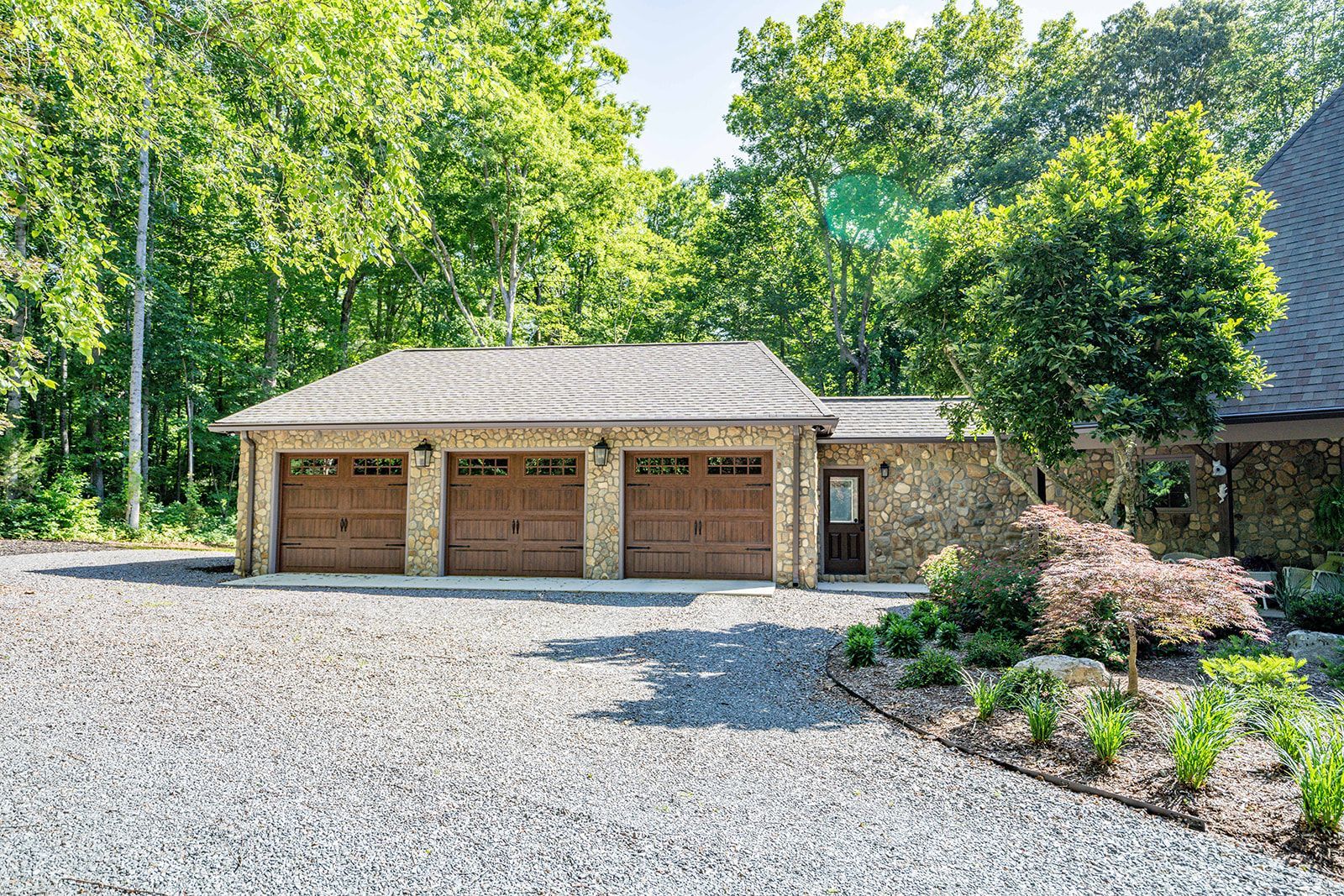 A large stone garage with three garage doors is surrounded by trees and gravel.