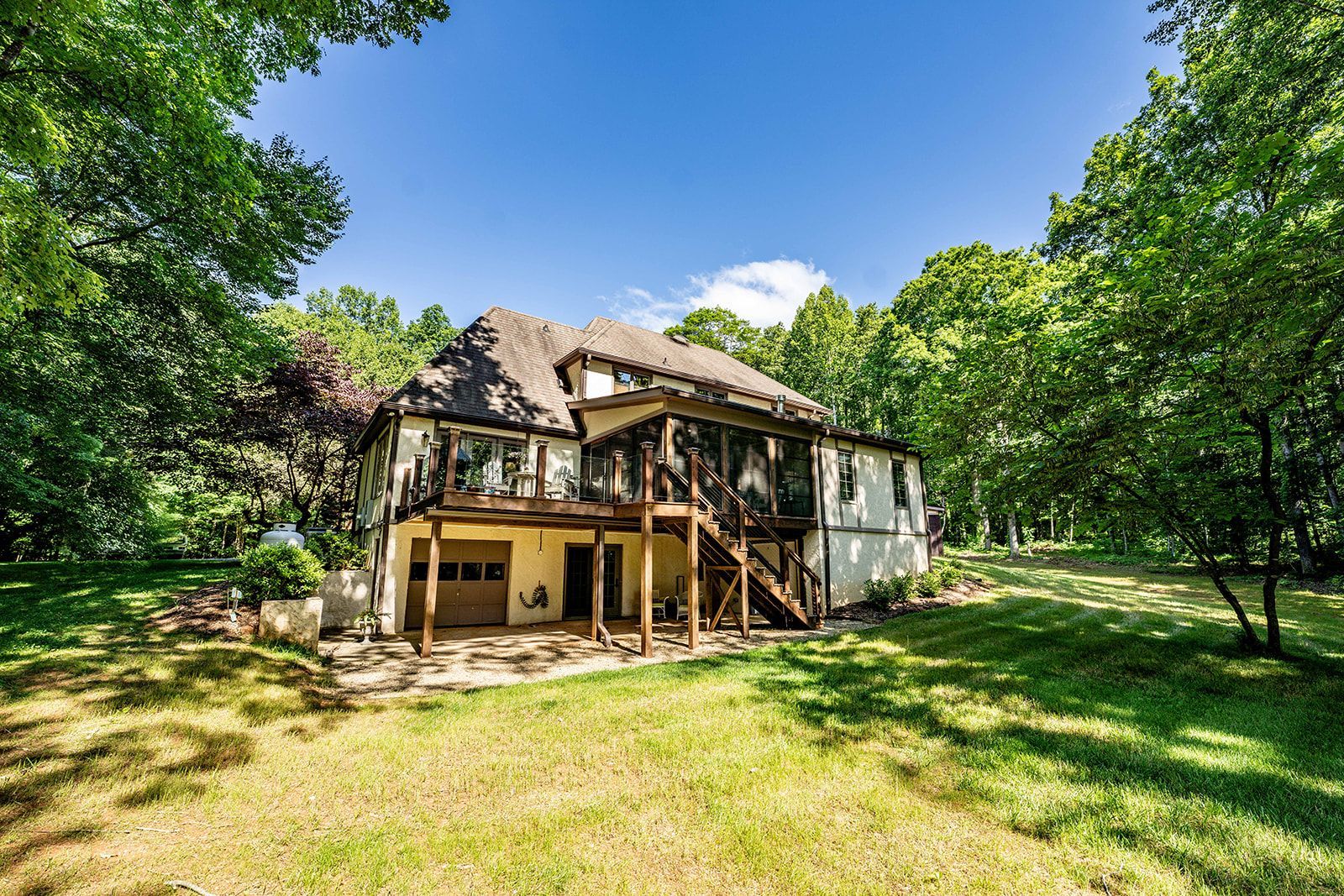 A large house with a deck and stairs in the middle of a lush green field surrounded by trees.