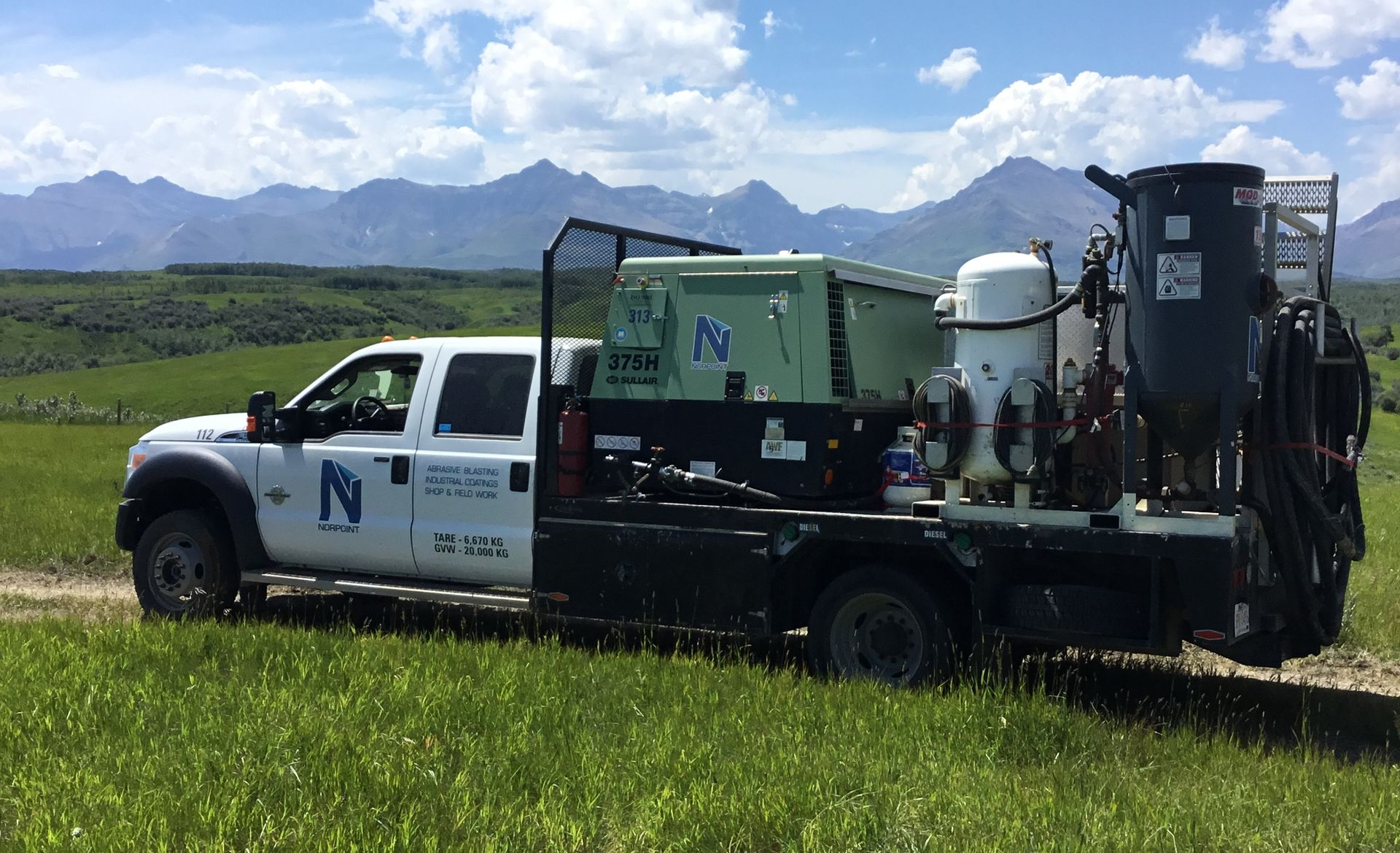 White utility truck with mounted equipment driving through a grassy field, mountains and clouds in the background