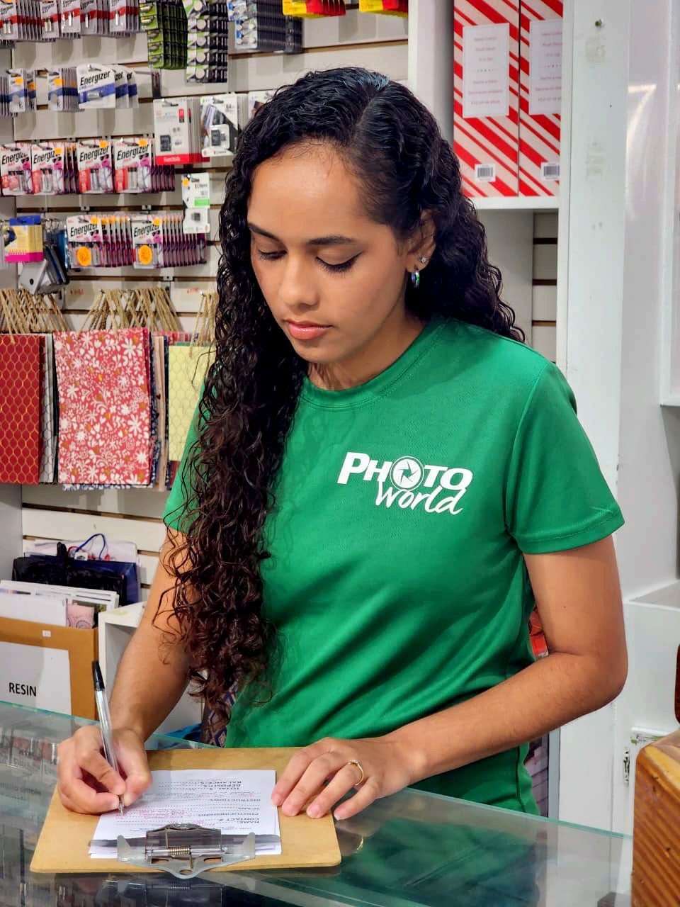 Woman in green Photo World shirt, writing on clipboard behind a counter in a store.