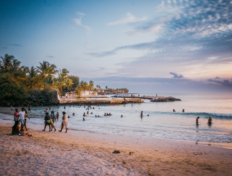 People swimming and walking on a beach with gentle waves under a colorful sunset sky.