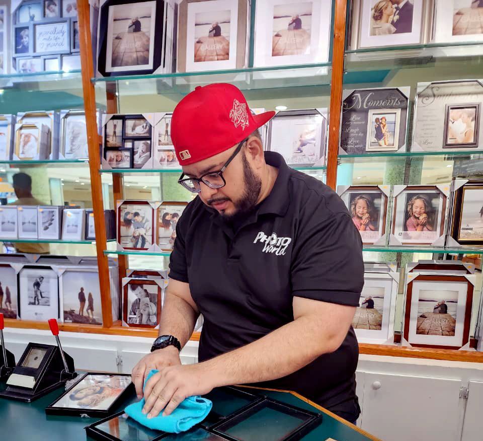 Man in red cap and black shirt cleaning picture frames at a store counter.