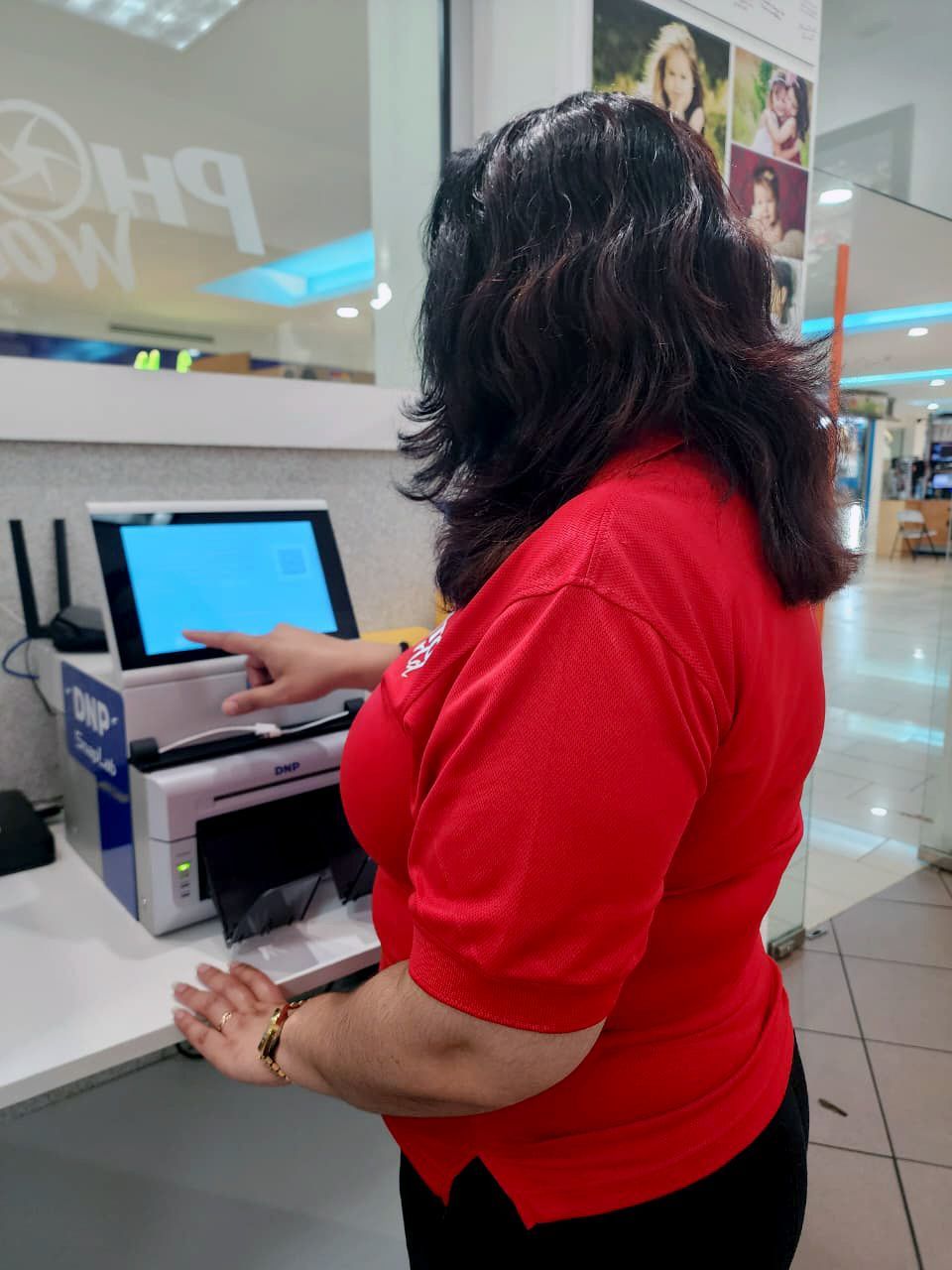 Woman in red shirt points to a photo printing kiosk screen in a store.