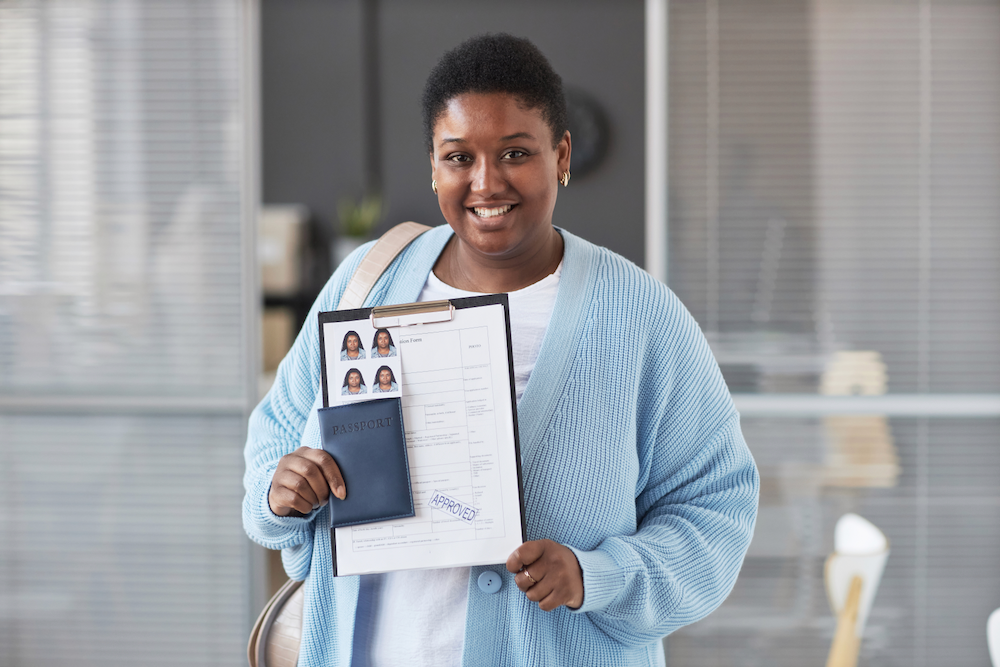 Black woman smiling, holding passport, photos, and documents, in a light blue cardigan.