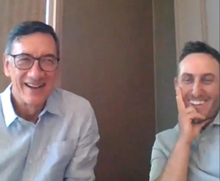Two men wearing glasses are sitting next to each other and smiling.