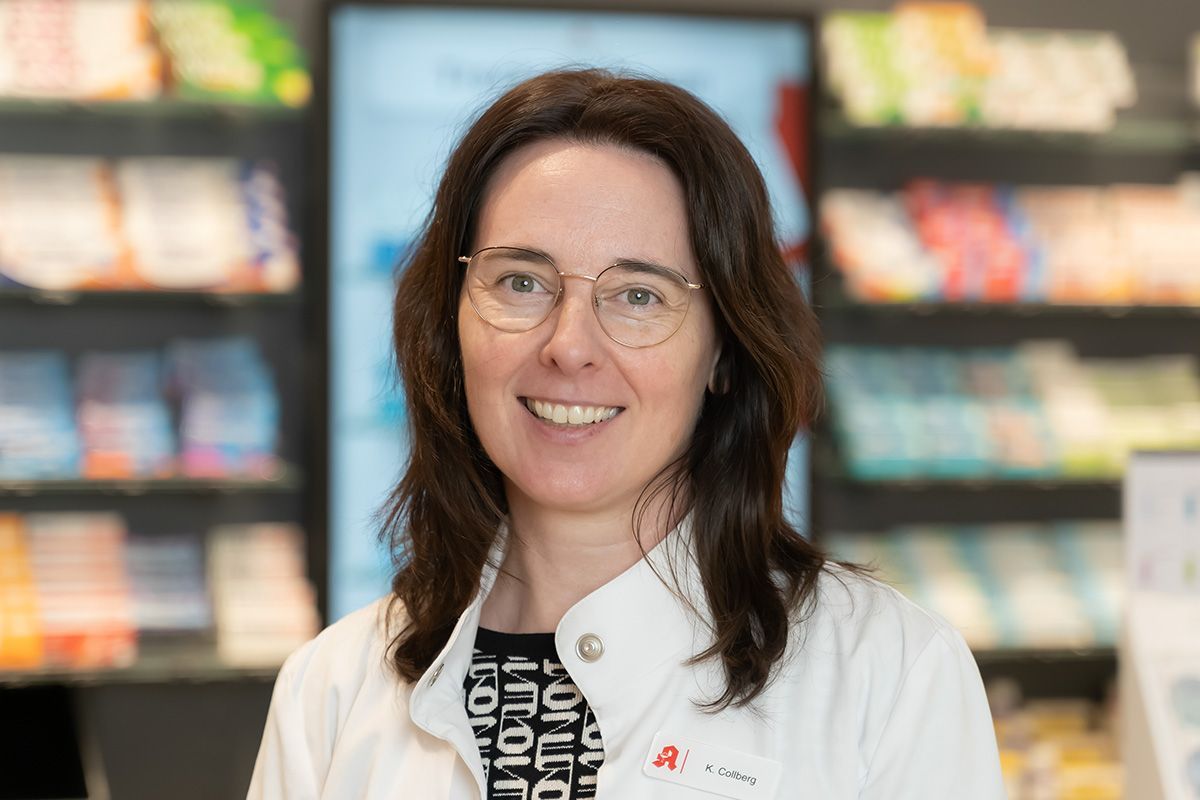 Eine Frau in einem weißen Kittel und mit Brille lächelt in einer Apotheke.