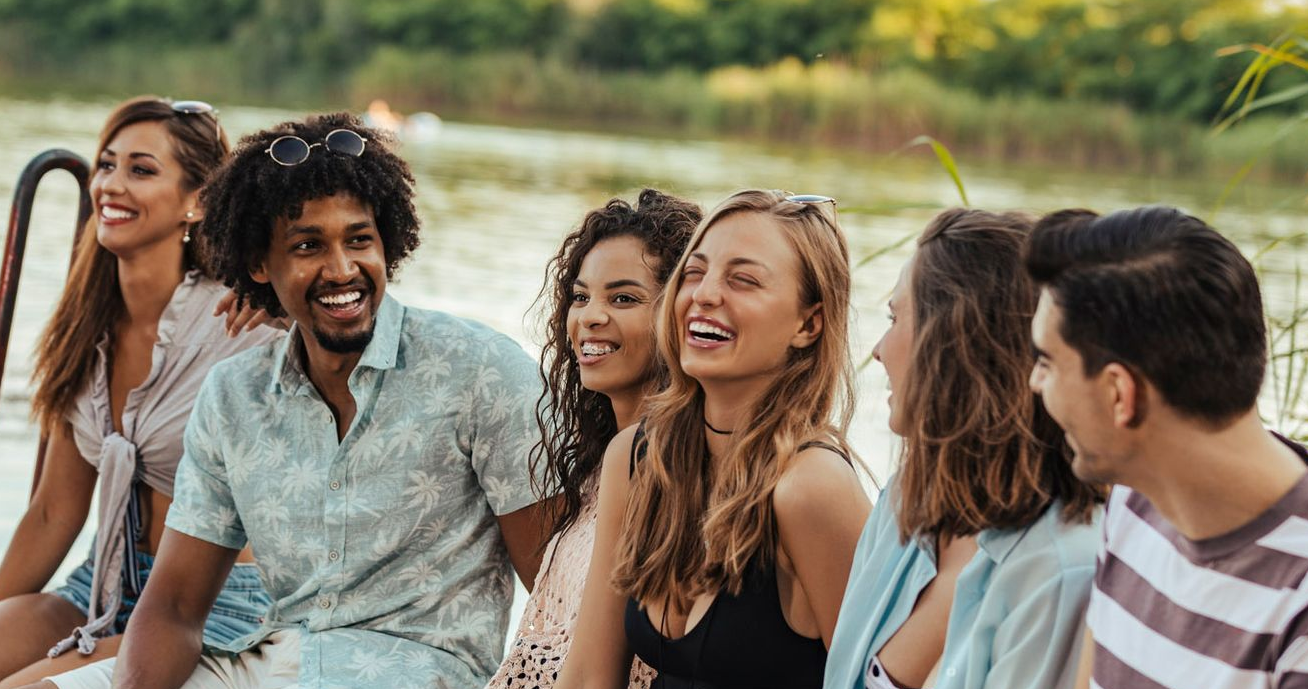 Five friends smiling and chatting on a boat by a lakeshore at sunset.