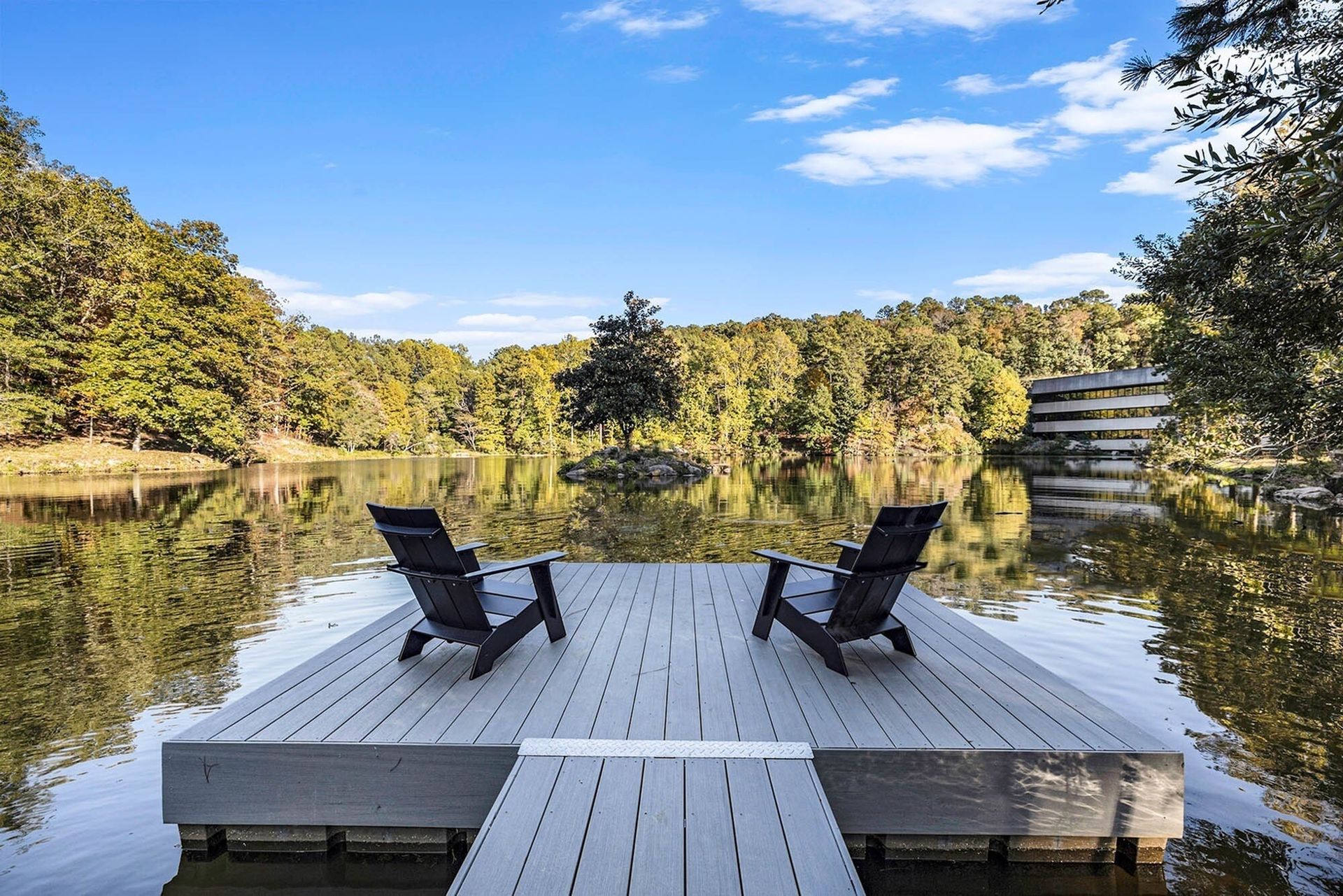 Two Adirondack chairs on a dock overlooking a lake surrounded by trees; sunny day.