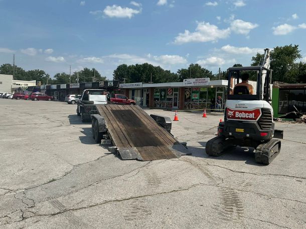 Bobcat mini skid steer near a loading ramp in a gravel lot beside shops under a blue sky