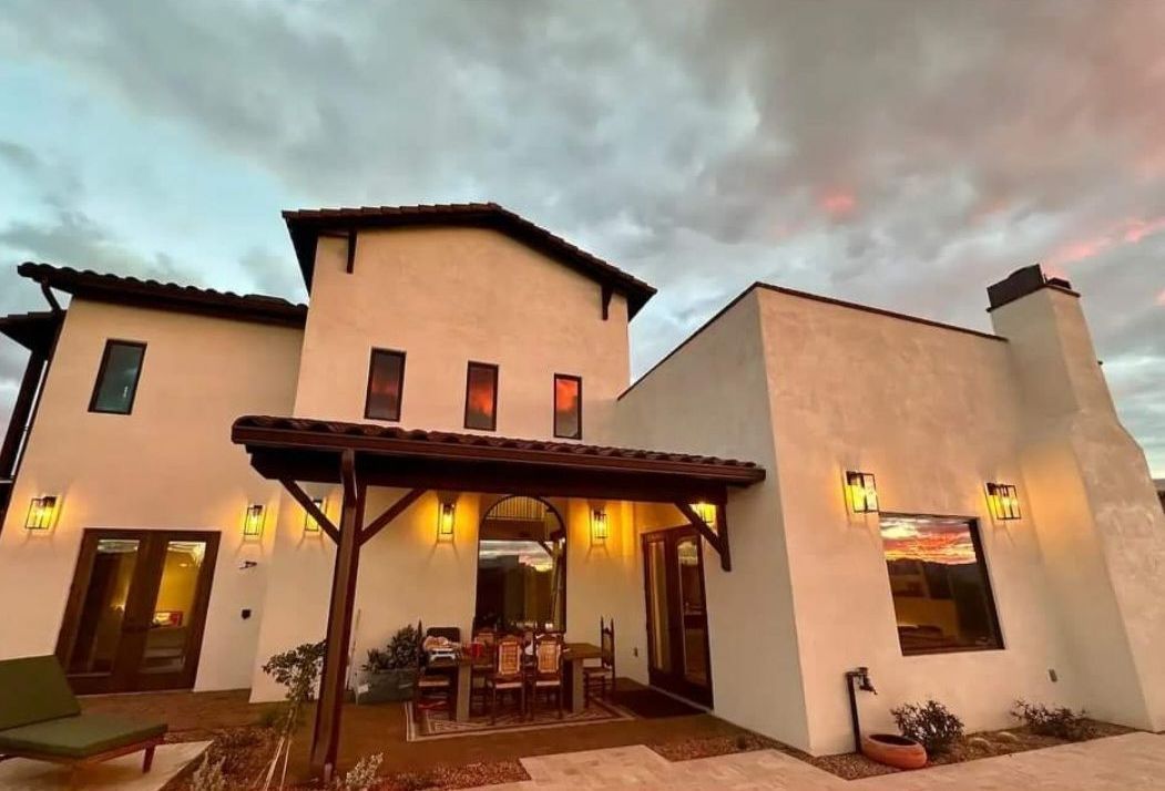 Exterior of a stucco house with a covered patio, dining table, and sunset sky.